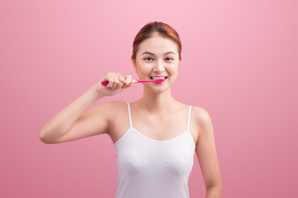 Smiling young woman with healthy teeth holding a tooth brush over pink background.